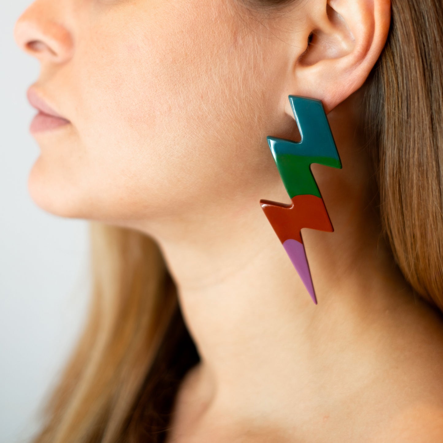 Brinco de resina em formato de raio nas cores petróleo, verde, terracota e violeta, usado por uma pessoa para demonstração.