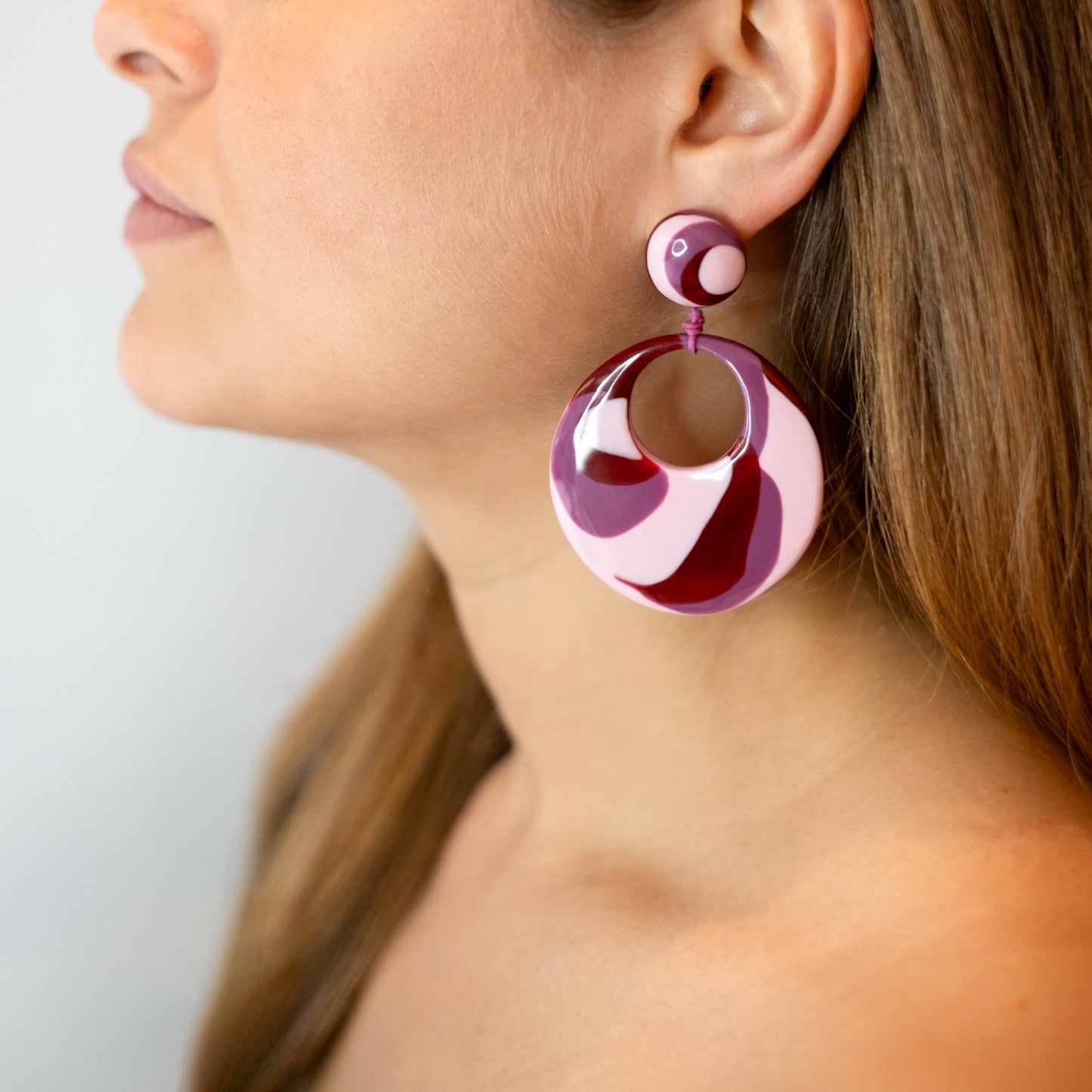 Brinco de resina em formato redondo nas cores vermelha, rosa claro e violeta, sado por uma pessoa para demonstração.